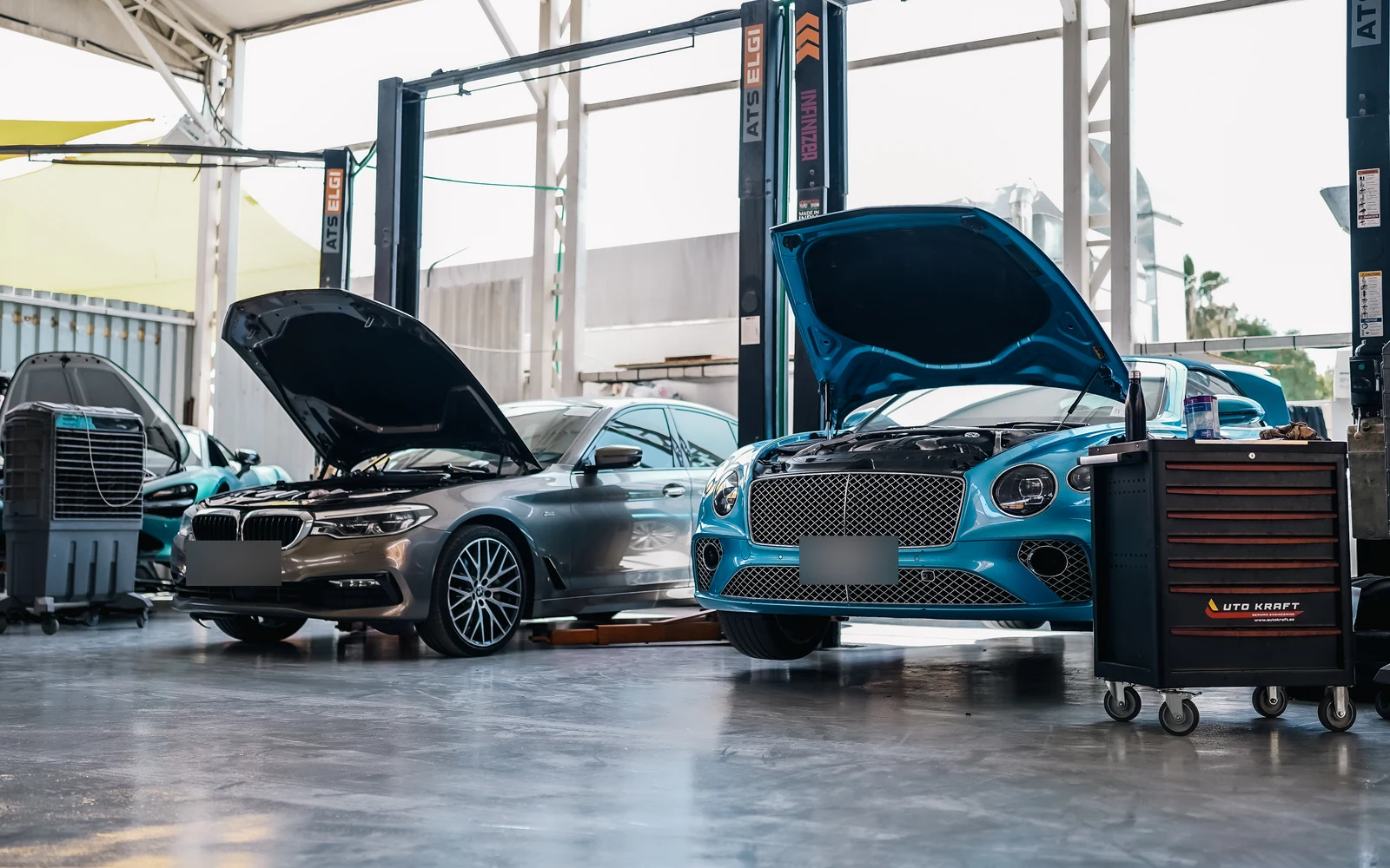 A turquoise Bentley beside a BMW, both with their bonnets open in the workshop.
