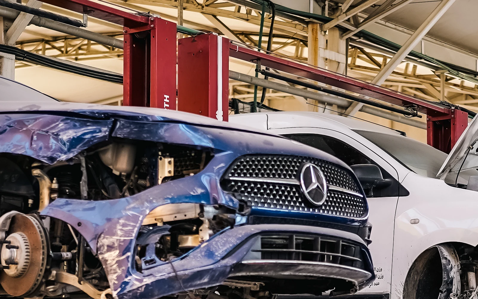 A blue Mercedes with a damaged front bumper and headlamp area on a workshop lift.